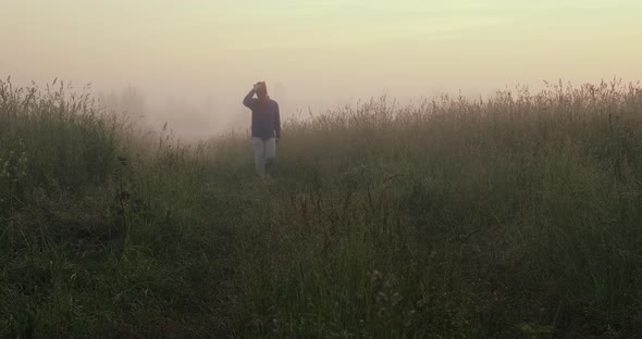Young Man in Cowboy Hat Walks Through a Field in Fog and Admires the Rising Sun