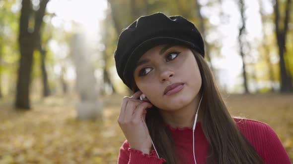 Girl Looking Admiringly in Autumn Park While Pushing the Earphones to Her Ears alt