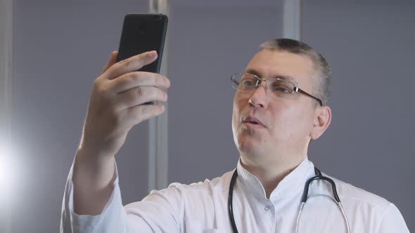 male doctor smiles and grimaces while taking a selfie on his phone alt