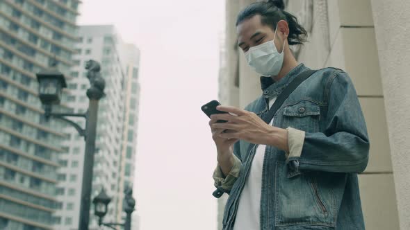 Asian man wearing a mask using a smartphone while standing on the street in the city.