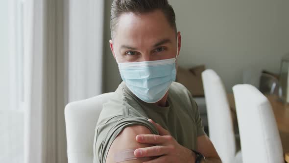 Portrait of caucasian man wearing face mask showing his vaccinated shoulder at home alt