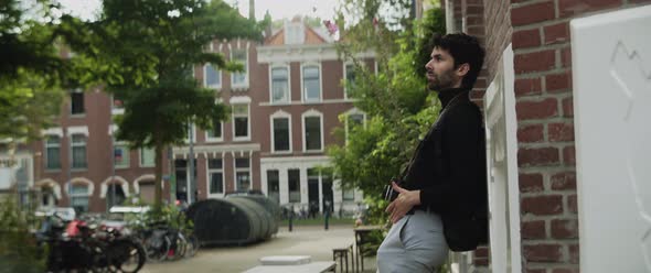 Caucasian man with beard and photocamera leaning up against a building in the city alt