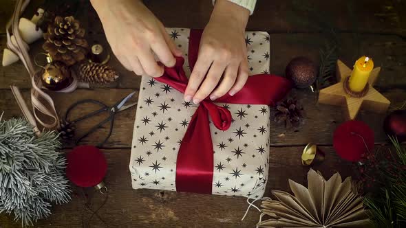 Hands wrapping stylish Christmas gift with red ribbon on rustic wooden table alt
