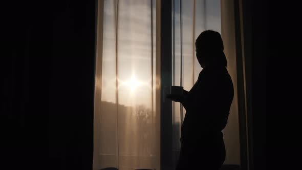 The Silhouette of a Woman at the Window Drinks a Hot Drink and Looks at the Rising Sun alt