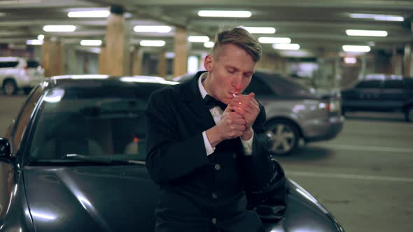 Young Man in a Black Suit with a Bowtie Smoking a Cigarette Sitting at the Bonnet of the Black Car alt