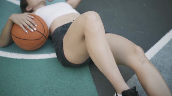 Camera Moves Along Fit Young Caucasian Woman Lying on Basketball Sports Ground with Orange Ball and alt