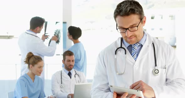 Doctor Using Tablet While Staff are Working Behind Him alt