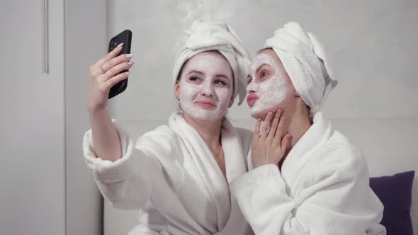 Two Girls in Bathrobes and with Clay Masks on Their Faces Take Selfies