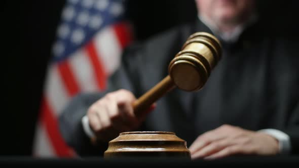 Judge's Hand Banging A Gavel On Block Against American Flag In United States Court alt