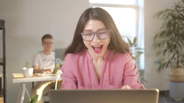 Beautiful Asian Businesswoman in Glasses Sitting at Her Office Desk in Front of Laptop Raising Arms alt