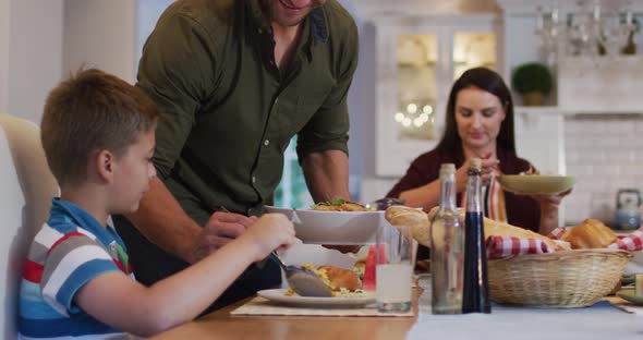 Smiling caucasian father standing at table serving son food before family meal alt