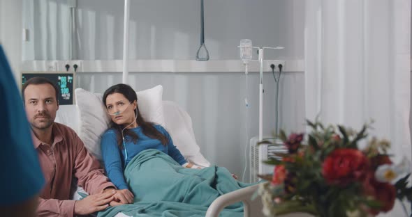 Sick Woman Patient Lying in Bed and Husband Sitting Near Listening To Doctor in Hospital Ward alt