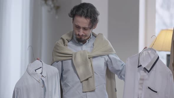 Dissatisfied Male Dressmaker Holding Two Shirts on Hangers Looking at Camera Shaking Head No alt