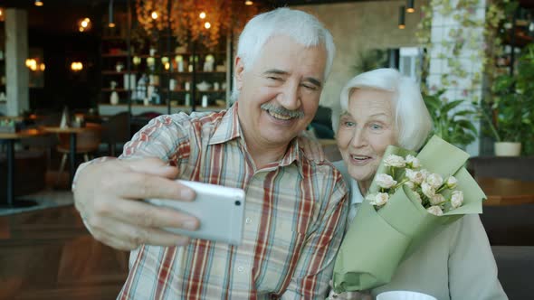 Beautiful Elderly Couple Taking Selfie in Cafe Using Smartphone Holding Flowers alt