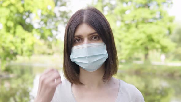 A Young Caucasian Woman in a Face Mask Looks at the Camera in a Park  Closeup alt