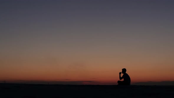Guy Sits on the Sand and Smokes an Electronic Cigarette Against the Sunset. Silhouette alt