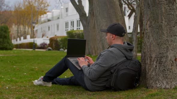 Young Man is Sitting in a Park in a Tourist Place Working on a Laptop alt