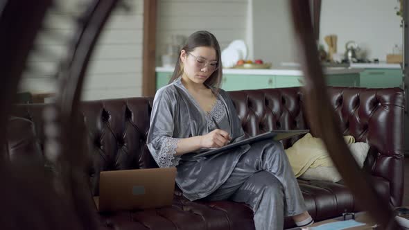 Talented Young Slim Asian Woman in Pajamas and Eyeglasses Drawing Sketch on Graphics Tablet Sitting alt