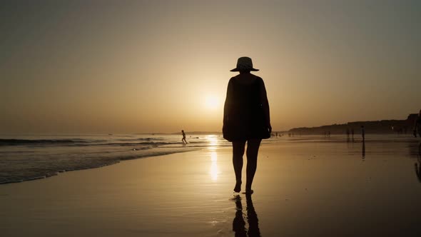 The Silhouette of a Woman in a Summer Dress and Panama Walks Along the Beach at Low Tide at Sunset