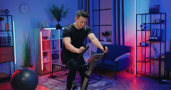 Asian Guy in Sportswear Exercising on Stationary Bike During Evening Home workout alt