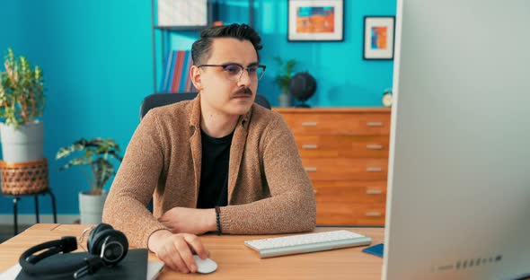 A Young Smiling Businessman Works in an Office on Computer Holds Mouse in Hand and alt
