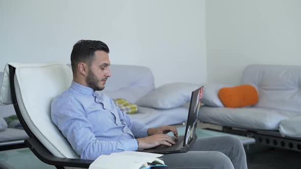 Young Bearded Businessman Sitting in Home Office at Table, Writing Documents and Working on Laptop alt