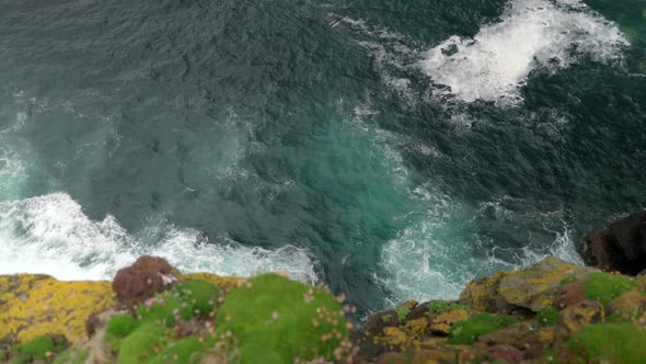 Waves gently crash over rocks and against the base of a sea cliff in the deep teal coloured ocean wh alt
