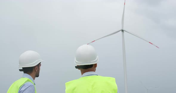 Engineers in white helmets stand and look at wind turbine.