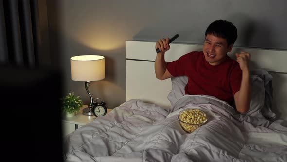 cheerful young man watching sport TV and win a game with arm raised on a bed at night