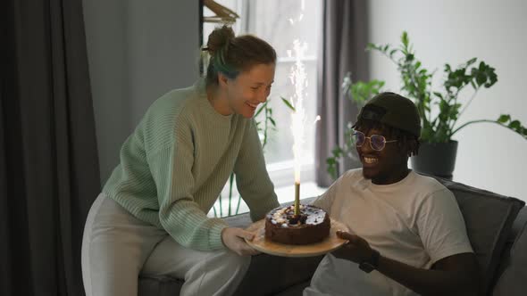 Young Wife Brought the Birthday Cake with Big Sparkling Candle to Her Husband alt
