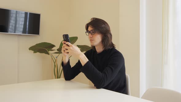 Young man with glasses in a beige room typing on his cell phone, sitting at a table alt