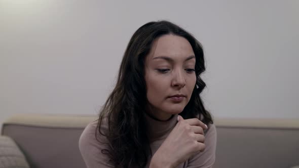 Portrait of a Darkhaired Woman Pensive in a Light Jacket Sitting on a Sofa in a Bright Room alt