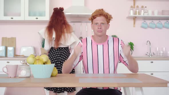 Portrait of Hungry Redhead Man Sitting at the Table in Kitchen Gesturing As Woman Cooking  alt