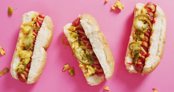 Video of hot dogs with mustard, ketchup and jalapeno on a pink surface alt