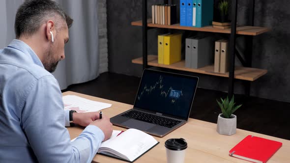 Man Trader Broker Uses Laptop with Stock Exchange Chart Makes Notes in Notebook alt