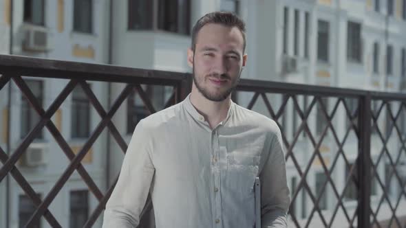 Portrait of a Pretty Young Man on the Terrace Looking in the Camera Holding Laptop in Hands alt