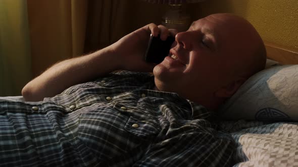 Man Smiling Lying in Bed Talking on Smartphone alt