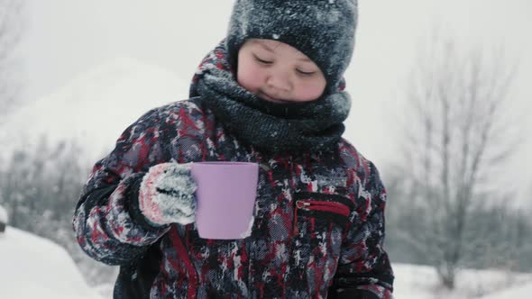 Cheerful boy drinking hot tea from cup in snowy forest at winter walk alt