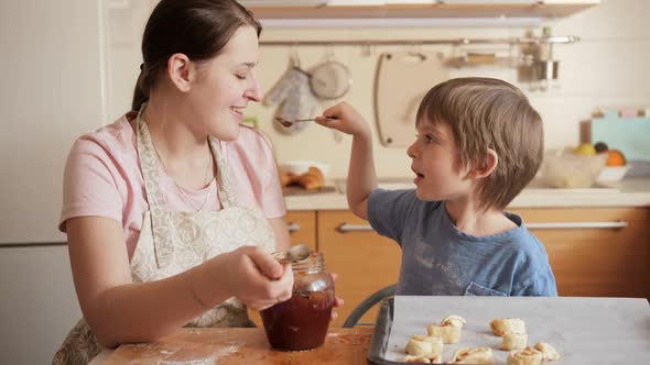 Smiling Little Boy Feeding His Mother with Jam From Spoon While Making Biscuits at Home alt