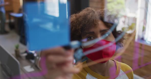 African american woman wearing face mask writing on glass board at modern office alt