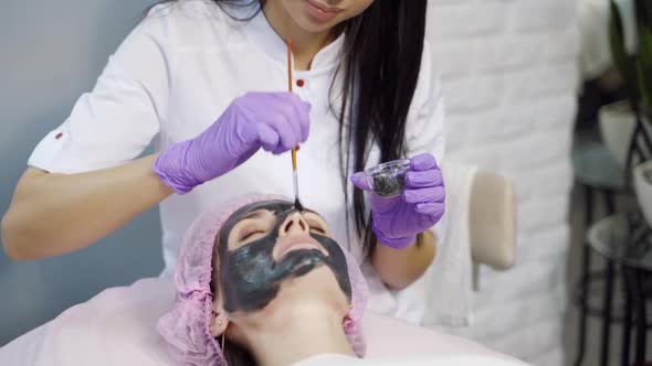 An experienced cosmetologist carefully applies a black cleansing mask to a woman’s face alt