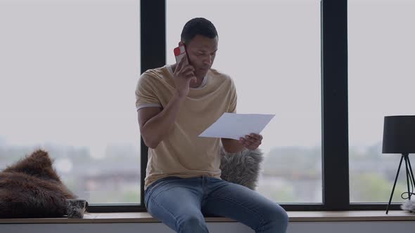 Portrait of African American Male Analyst Examining Graph Talking on the Phone Sitting on Windowsill alt