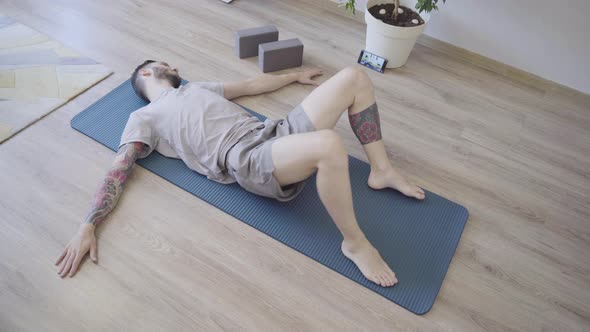 Young Man Relaxing and Doing Reclined Spinal Twist Yoga Pose at Home alt