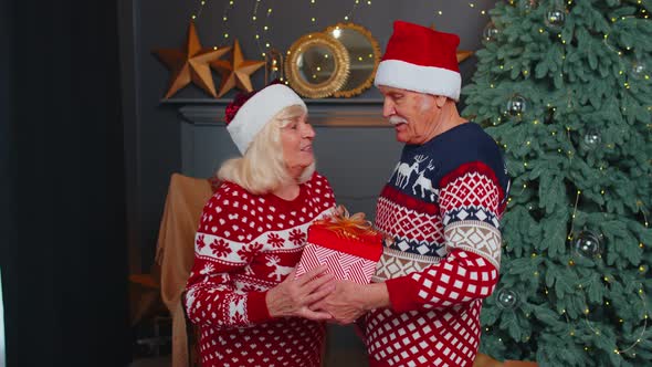 Grandfather Gifting Christmas Present Box to Surprised Grandmother Senior Couple in Santa Claus Hats alt