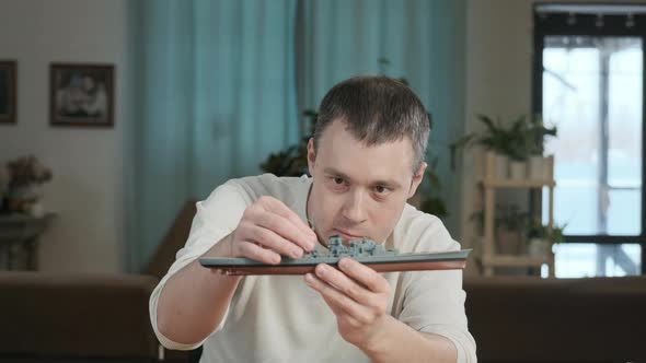 male in  room at table builds a model of toy and carefully examines ship alt
