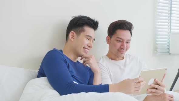 Asian handsome gay couple sit on bed using tablet in bedroom at home. alt