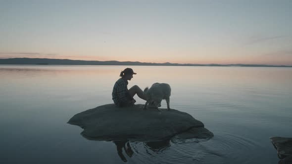 Woman and Dog Sitting on Rock in Lake at Sunset alt