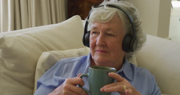 Caucasian senior woman wearing headphones listening to music while holding coffee cup at home alt