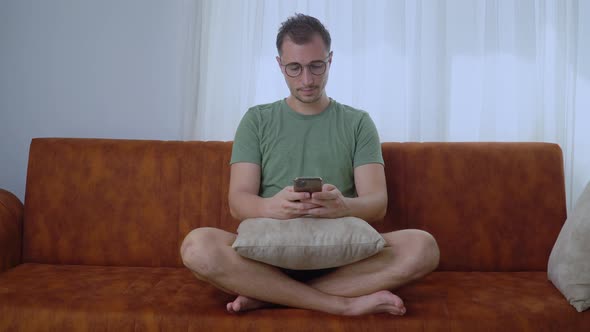 A Man with a Phone in the Lotus Position Sits on a Sofa and Corresponds in a Smartphone