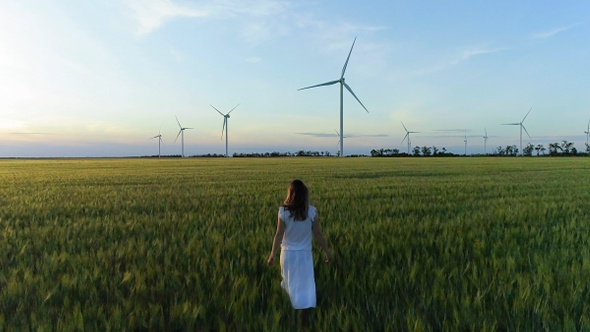 Beautiful girl walking on a green wheat field with windmills for electric power production alt